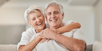 Alterer Mann sitzt lachend auf der Couch, während seine Frau ihn von hinten umarmt